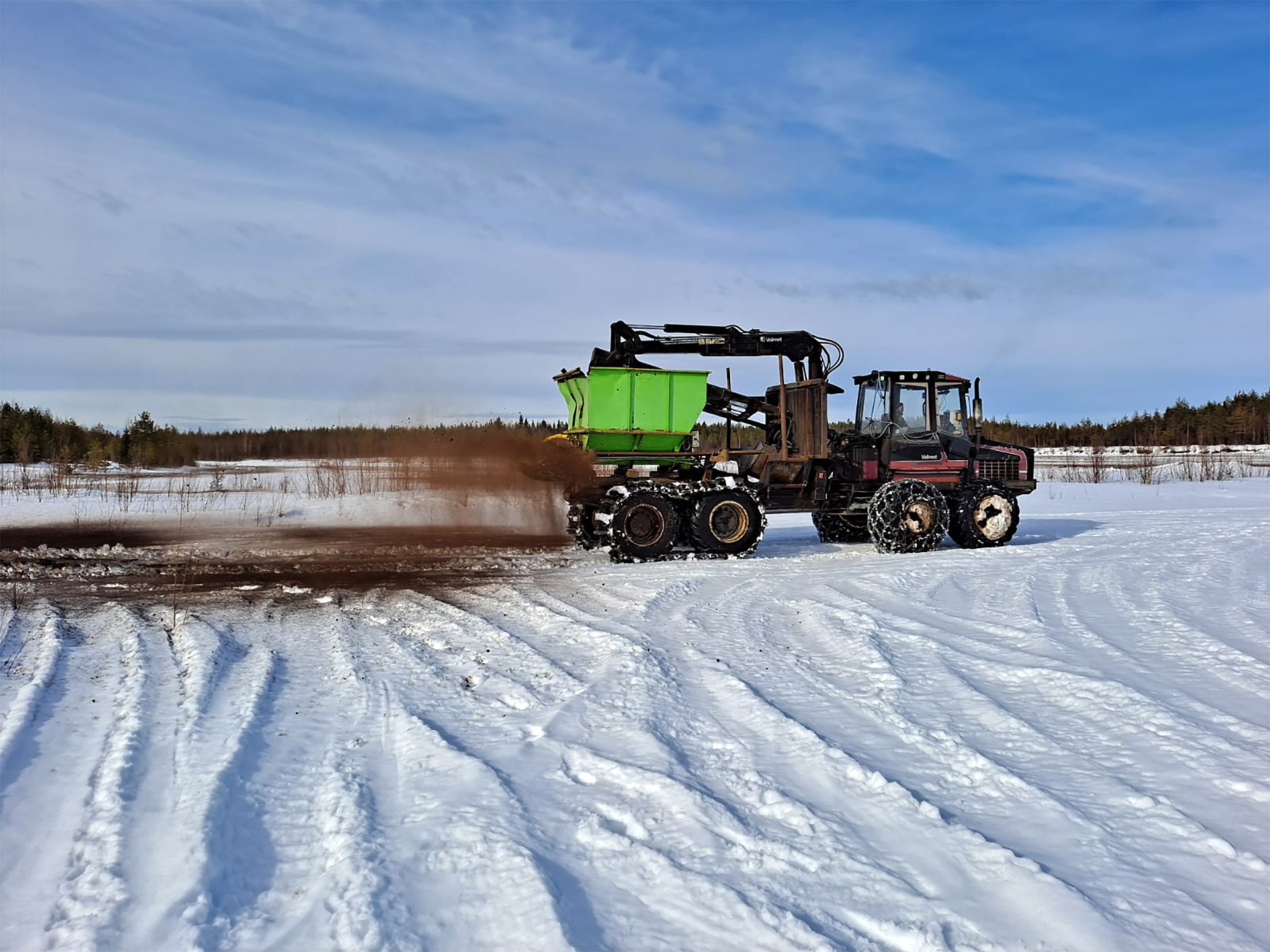Traktori etenee lumella ja peräkärrystä lentää hienoa ainesta ilmaan.