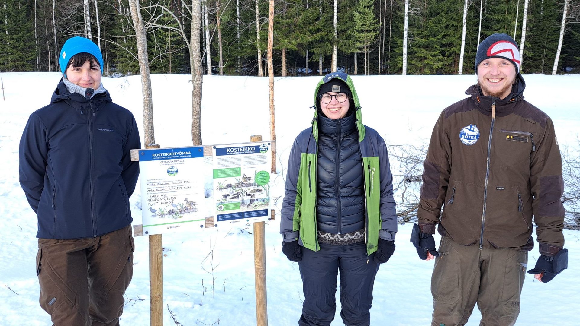 Tre personer står i ett vinterlandskap bredvid en skylt.
