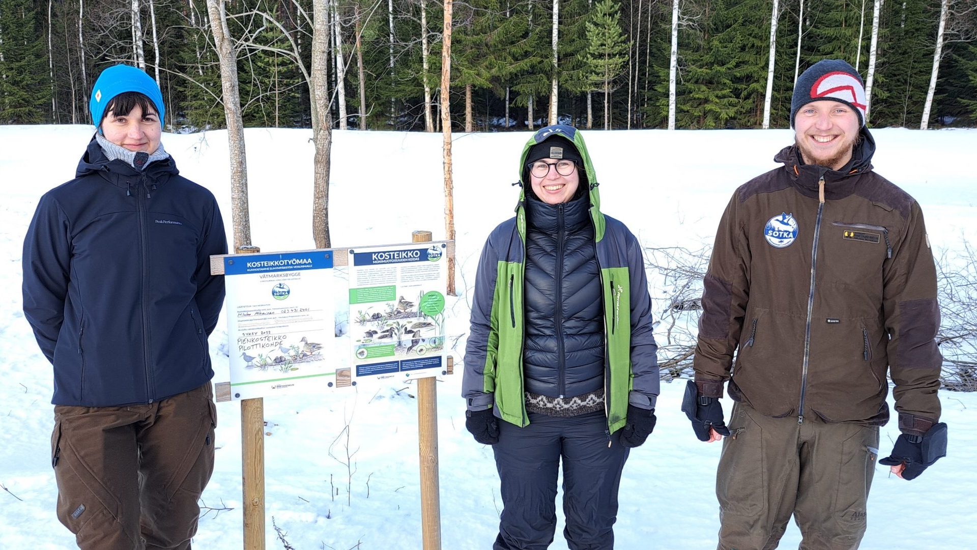 Tre personer står i ett vinterlandskap bredvid en skylt.