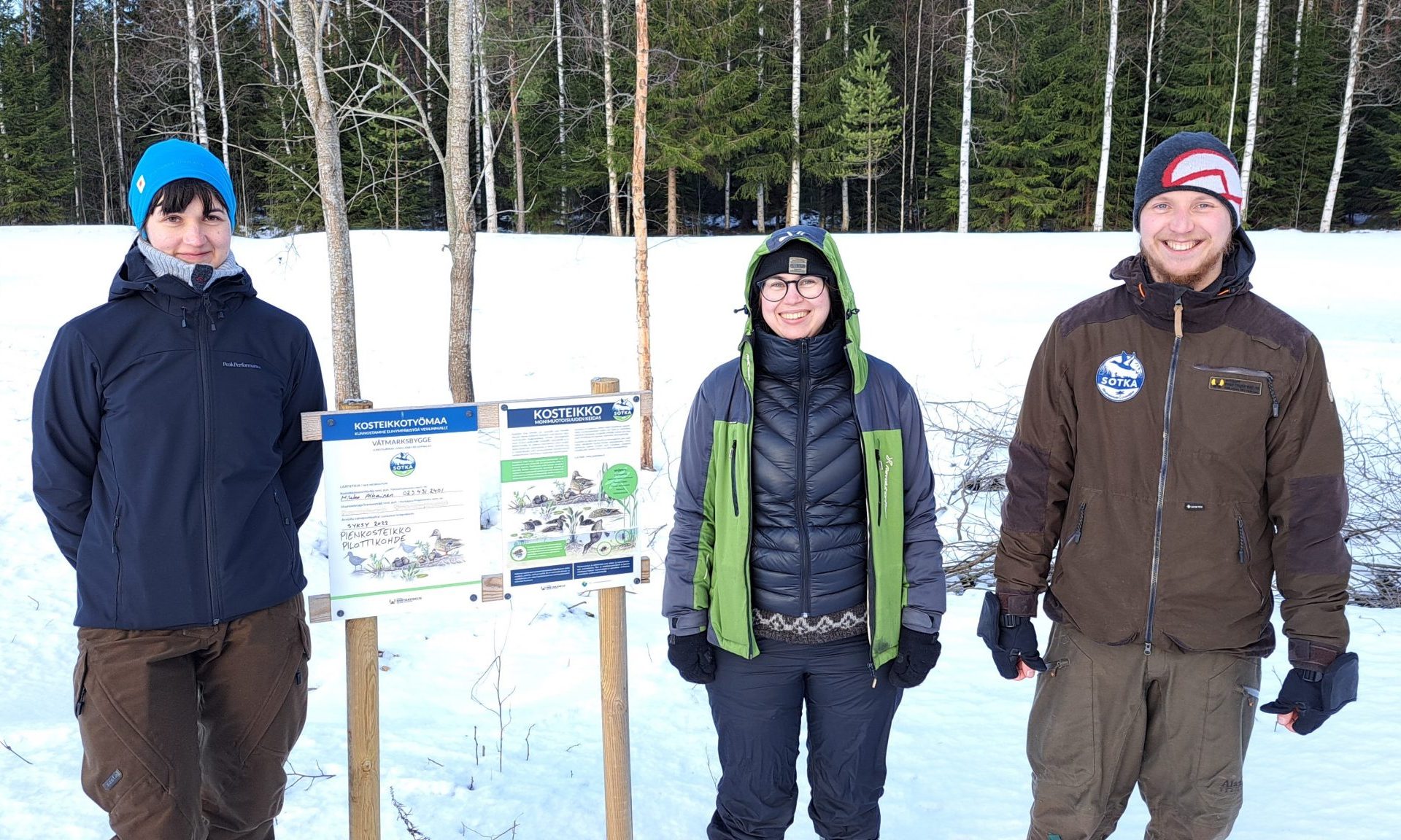 Kolme henkilöä seisoo talvisessa maisemassa kosteikko-kyltin vieressä.