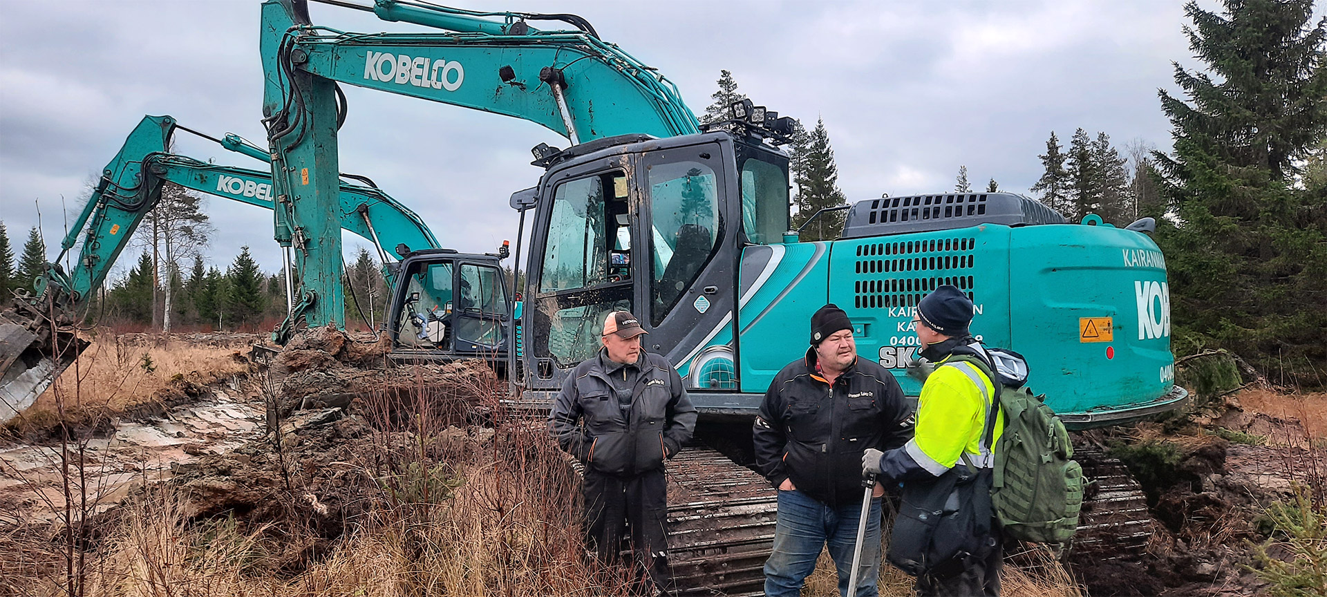 Två grävmaskiner som bearbetar ett våtmarksområde. I förgrunden står tre personer och diskuterar bredvid en av maskinerna.